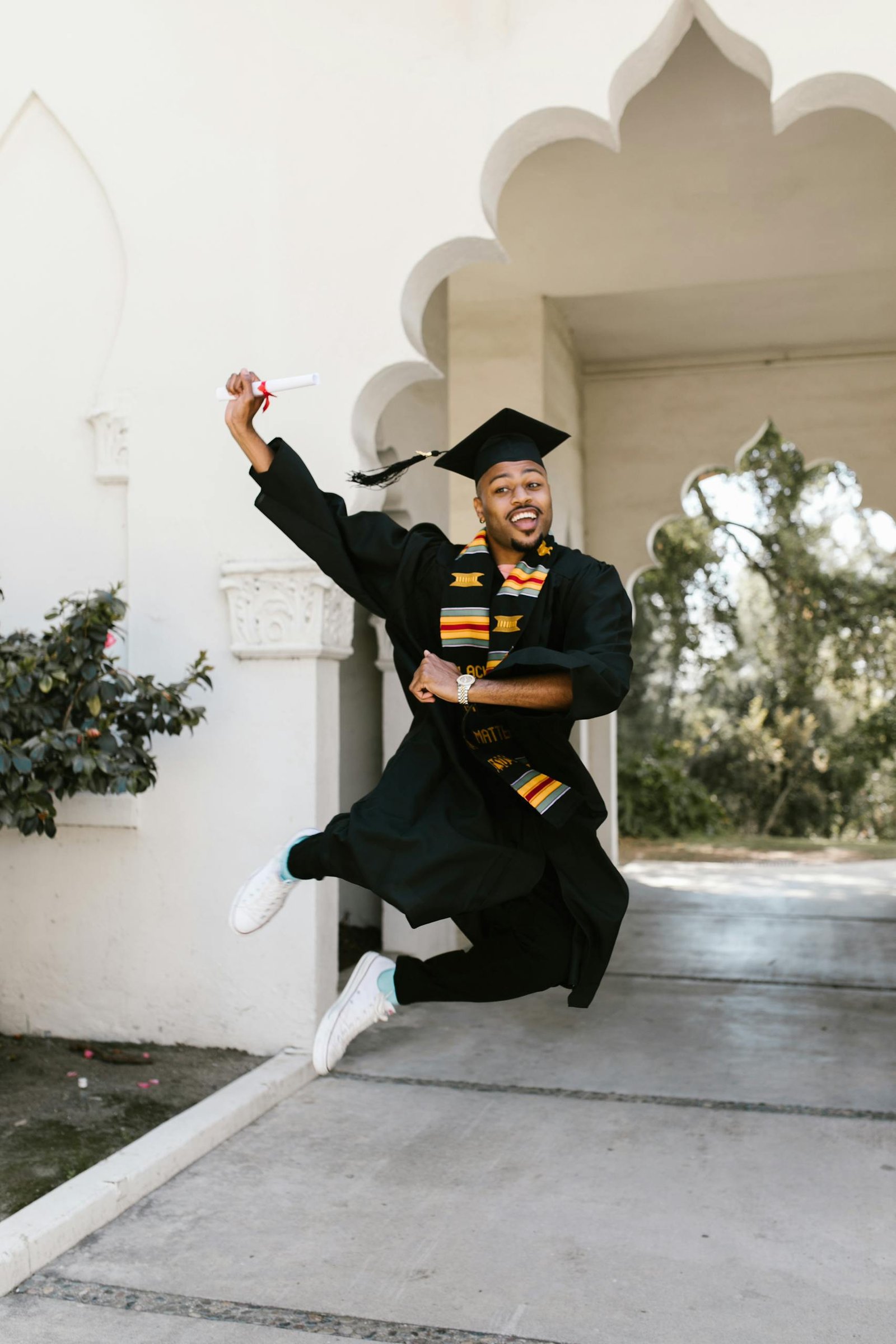 Celebratory graduate jumping in traditional attire, holding a diploma outdoors with a joyful expression.